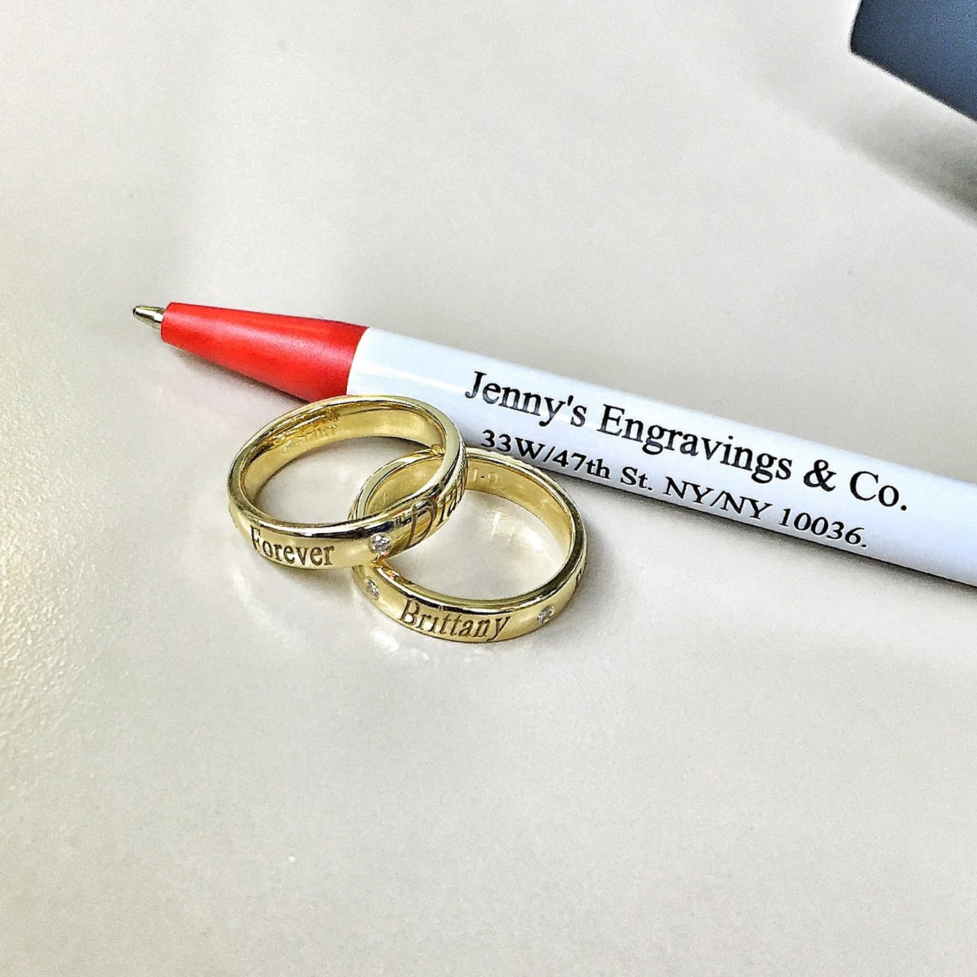 Two gold wedding bands beside a pen printed with the Jenny's Engraving details